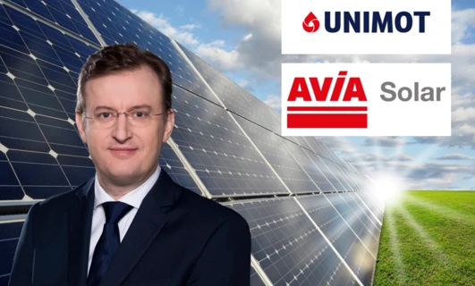Pracownik umysłowy w garniturze i krawacie stoi obok panelu słonecznego z reklamą UNIMOT, firmy specjalizującej się w energii słonecznej. W tle widoczne jest również AVİA Solar.