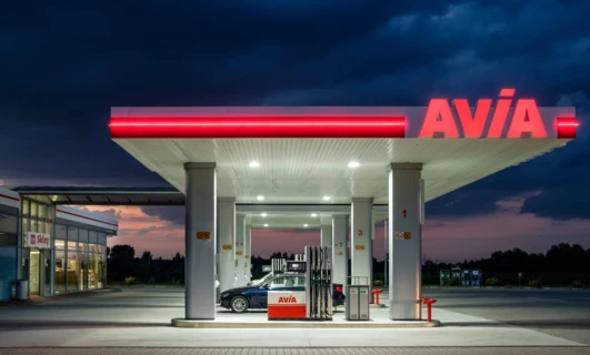 Obraz nowoczesnej stacji benzynowej z dystrybutorem paliwa i sklepem sprzedającym produkty, takie jak benzyna. Widoczne są wyraźnie oznaczenia marki AVIA.